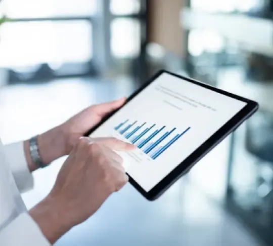 Person holding a tablet displaying a bar graph with blue columns and text, in a well-lit office setting.