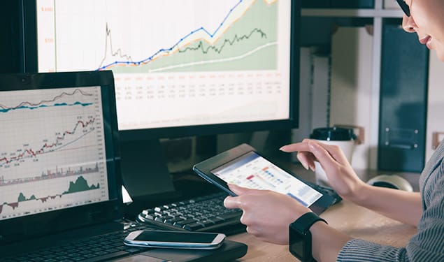 A person uses a tablet computer while sitting at a desk with multiple screens displaying financial graphs and charts.